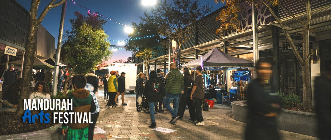 A large number of people, some in groups or pairs, walk through an evening market that is lit by festoon lights